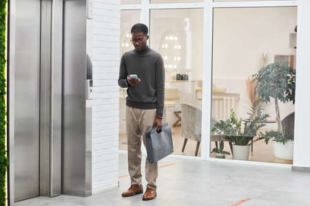 Minimal full length portrait of young black businessman standing by elevator in modern office...