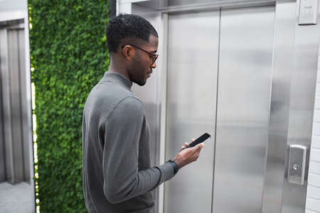 Minimal waist up portrait of young black businessman waiting for elevator in office building and...