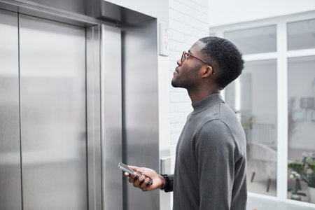 Minimal side view of young black businessman waiting for elevator in office building and holding...