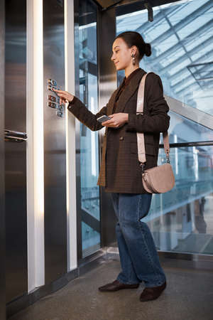 Full length side view at young professional woman pushing button in glass elevator in modern office...