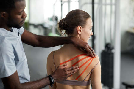 Close up rehabilitation therapist putting tape on shoulder of young woman in physiotherapy clinic,...