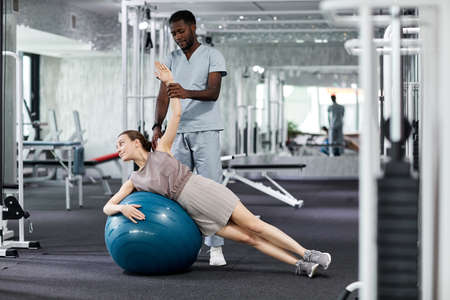 Portrait of young therapist assisting woman doing fitness exercises in rehabilitation clinic, copy...