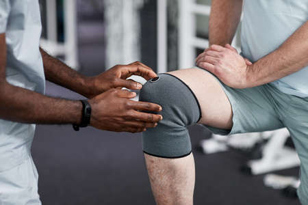 Close up of African American therapist examining senior man with knee injury in rehabilitation clinicの写真素材