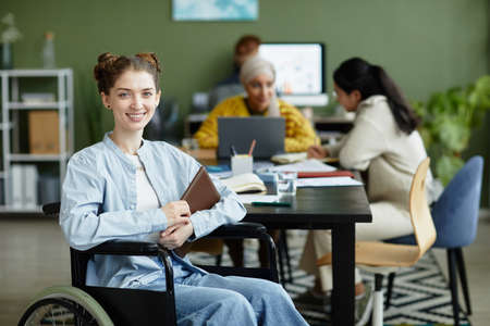 Portrait of smiling young woman in wheelchair looking at camera while working in office with diverse creative team, copy spaceの写真素材