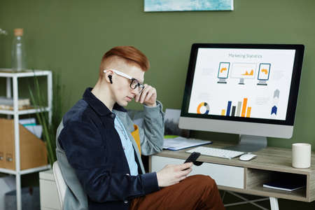 Side view portrait of trendy young businessman using smartphone while working at desk in green officeの写真素材