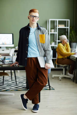 Full length portrait of red haired young man wearing vibrant style and smiling at camera while standing in officeの写真素材