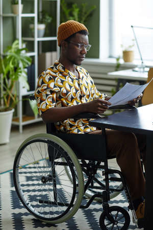 Full length portrait of black young man in wheelchair wearing creative style while working in officeの写真素材