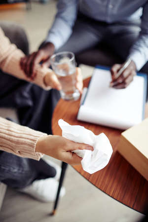 Psychologist offering glass of water and napkin to female patient crying in therapy session