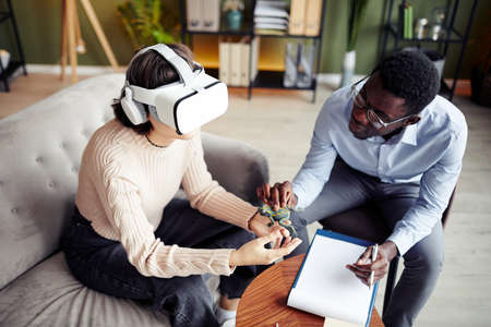 Therapist giving toy frog to young patient wearing virtual reality headset and asking to describe...