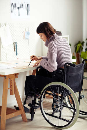 Creative young fashion designer in wheelchair checking sewing pattern she made for project