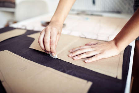 Hands of seamstress using chalk to outline cardboard sewing pattern on fabric when working in...