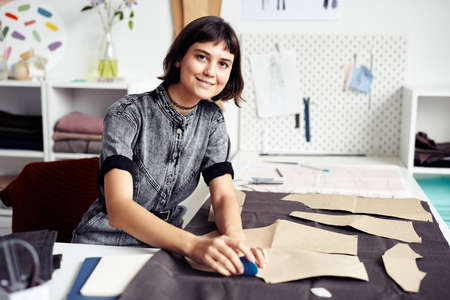 Smiling young seamstress outlining and cutting out garment details