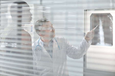 Through glass wall shot of doctor showing backbone x-ray image and explaining something to injured...