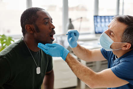 Young Black military officer sitting with mouth wide open doing PCR swab test for coronavirus in...