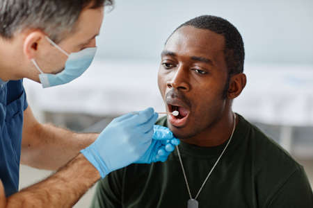 Unrecognizable mature doctor wearing mask and gloves taking mouth swab test from young African American army officerの写真素材