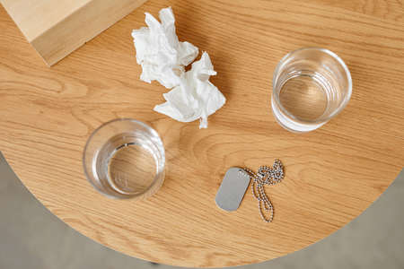 Top-down still life shot of used paper napkins, glasses with water and military dog tag on table,...