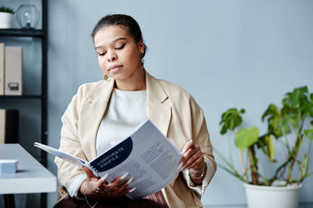 Minimal portrait of young black businesswoman reading report at workplace in office against grey...