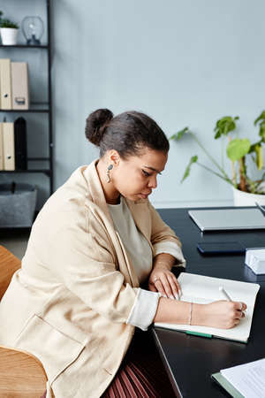 Vertical side view portrait of elegant black businesswoman writing in notebook while sitting at desk...