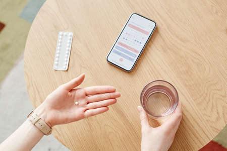 Top view closeup of unrecognizable woman taking birth control pills with glass of water