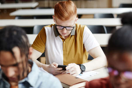 High angle portrait of red haired young man using smartphone at desk in college classroomの写真素材