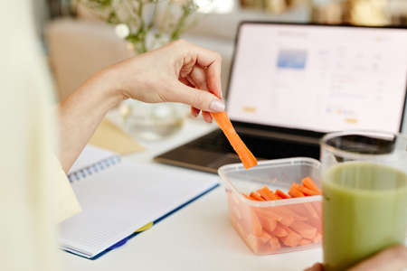 Unrecognizable woman working on laptop in office and eating fresh sweet carrot sticks with green...