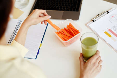 High angle view of unrecognizable woman working in office having fresh carrot sticks and vegetable...