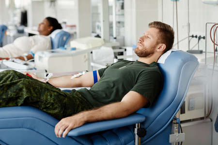 Side view portrait of military man donating blood while laying in chair at plasma donation centerの写真素材
