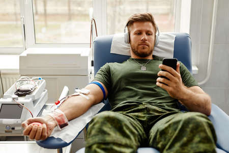 Portrait of young military man donating blood with comfort and watching videos at smartphone, copy spaceの写真素材