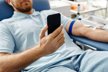 Close up of young man donating blood while lying in comfortable chair at med center and holding smartphoneの写真素材