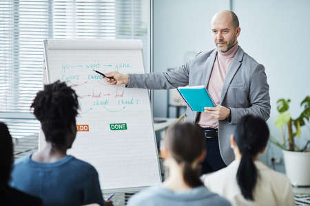 Portrait of mature man giving English lessons in office setting to group of people and pointing at whiteboard, copy spaceの写真素材
