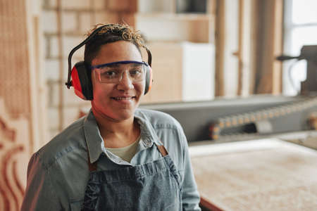 Portrait of female worker smiling at camera while wearing noise cancelling headphones in workshop,...