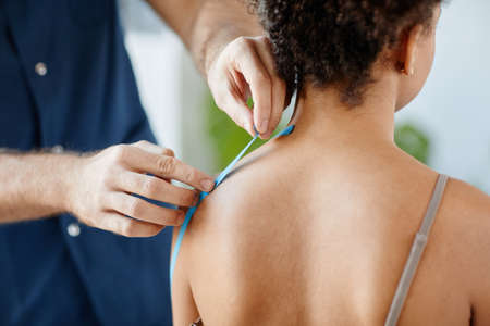 Close up rehabilitation therapist putting physio tape on shoulder of young woman to relieve muscle...