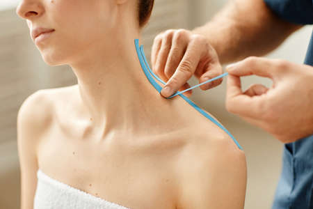 Close up of rehabilitation specialist putting physio tape on shoulder of young woman during therapy...