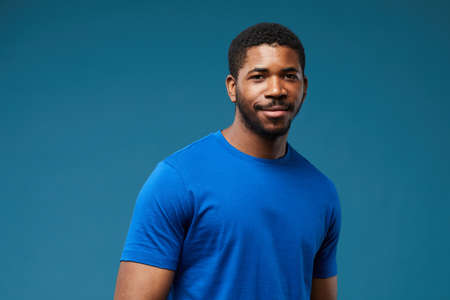 Portrait of smiling black man wearing blue shirt on vibrant blue background and looking at camera,...