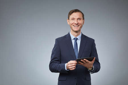 Waist up portrait of confident Caucasian businessman smiling to camera and holding digital tablet while standing against grey background, copy spaceの写真素材