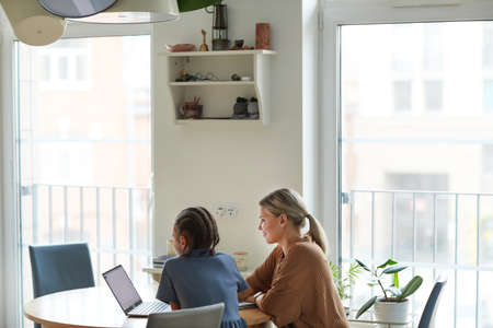 Minimal shot of multiethnic family, mother and daughter looking at laptop screen while studying together at home in minimal setting, copy spaceの写真素材