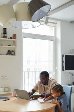 Vertical portrait of young black girl drawing at table with father working from home in cozy light...