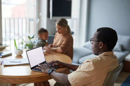 Side view portrait of black man using laptop at home with wife and children in background, copy...