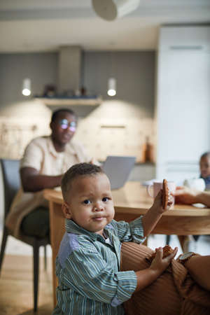 Vertical portrait of cute little boy looking at camera while eating cookie in kitchen with family in backgroundの写真素材