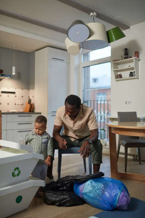 Vertical portrait of caring black father with baby son sorting plastic waste in kitchen