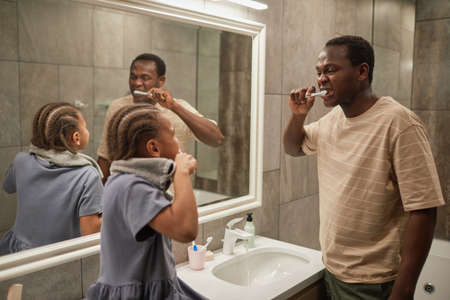Side view portrait of cute black girl brushing teeth with dad and laughing happily in bathroomの写真素材