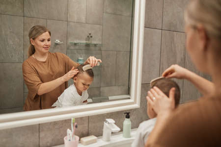Portrait of caring Caucasian mother combing hair or mixedrace baby boy by mirror in bathroom, copy...