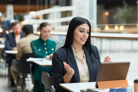 Portrait of young Middle Eastern businesswoman talking in video call meeting while sitting at table...