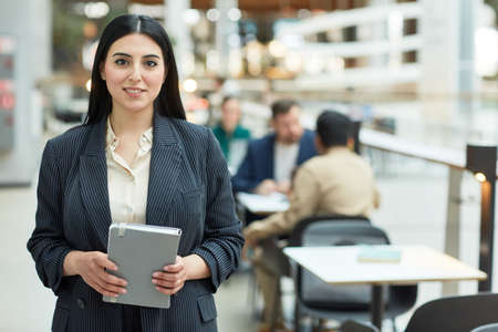 Waist up portrait of young Middle Eastern businesswoman holding planner and smiling at camera in modern office building interior, copy spaceの写真素材