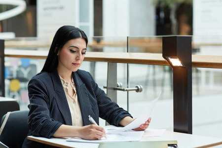 Portrait of young Middle Eastern businesswoman writing in document while working at cafe table in office building setting, copy spaceの写真素材