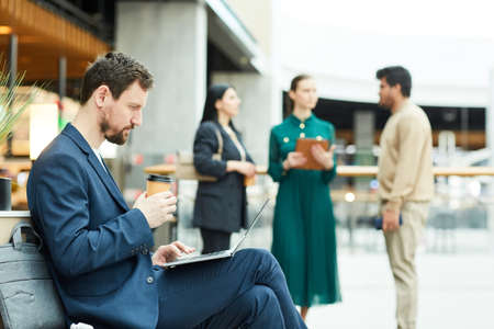 Side view portrait of bearded businessman using laptop and enjoying coffee while taking break in lounge area of office building, copy spaceの写真素材