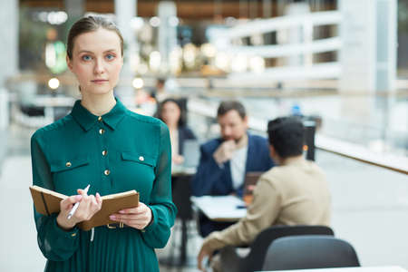 Waist up portrait of young businesswoman holding planner and looking at camera in office building...