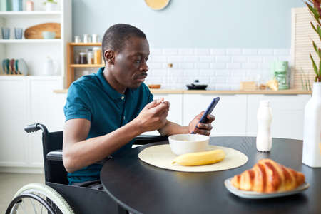 Portrait of black adult man with disability eating breakfast in home kitchen and using phone, copy...