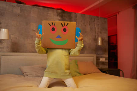 Portrait of unrecognizable carefree child playing with cardboard box and jumping on bed at homeの写真素材