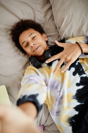 High angle POV shot of trendy black teenager wearing tie dye shirt while lying on bed and recording video for social mediaの写真素材